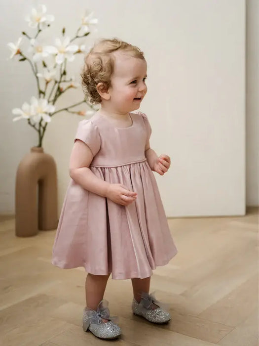 Teter Warm Harper pink baby party dress worn by a smiling toddler standing indoors with flowers in the background.