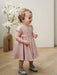 Teter Warm Harper pink baby party dress worn by a smiling toddler standing indoors with flowers in the background.