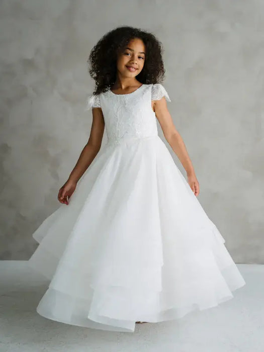 Teter Warm Lenni off-white communion dress shown on a smiling girl standing against a neutral background.
