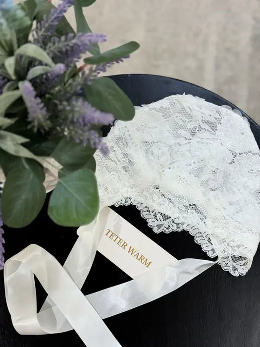 Teter Warm Leslie Lace Baby Bonnet displayed on a dark surface with white ribbons and flowers nearby.