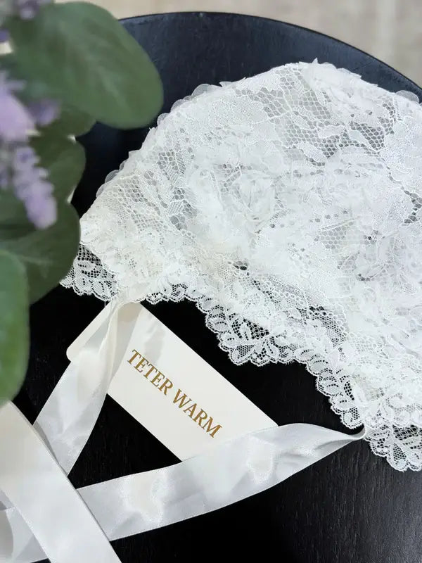 Teter Warm Leslie lace baby bonnet in white shown resting on a flat black surface with ribbon ties.