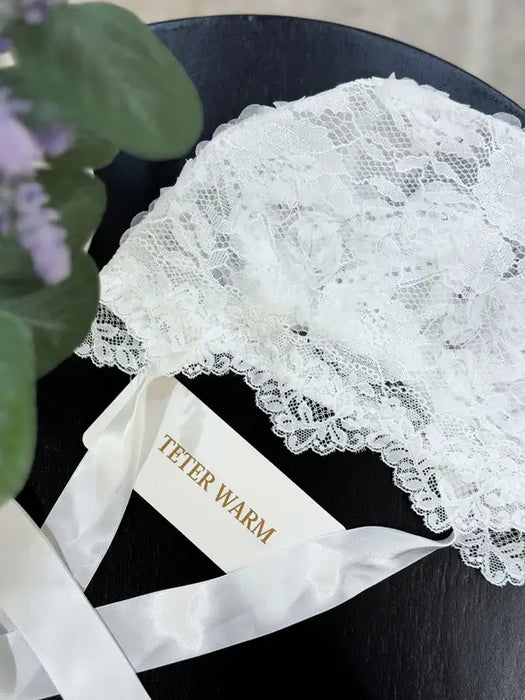 Teter Warm Leslie lace baby bonnet in white shown resting on a flat black surface with ribbon ties.