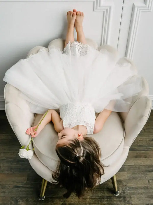 Off-white lace flower girl dress worn by a child lying playfully on a cream armchair holding a flower.