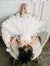Off-white lace flower girl dress worn by a child lying playfully on a cream armchair holding a flower.