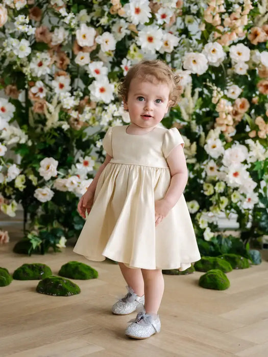 Teter Warm Melanie soft gold baby party dress worn by a toddler standing in front of floral backdrop.