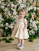 Teter Warm Melanie soft gold baby party dress worn by a toddler standing in front of floral backdrop.