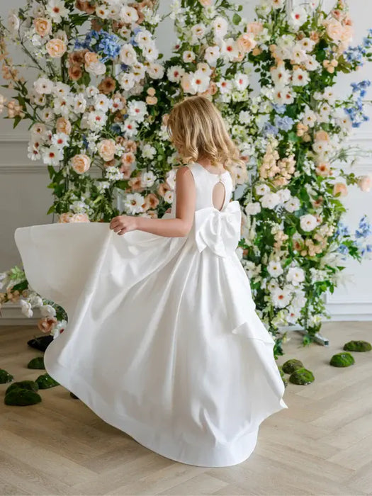 Teter Warm Penelope off-white communion dress with bow, worn by a girl in front of floral backdrop.