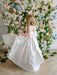 Teter Warm Penelope off-white communion dress with bow, worn by a girl in front of floral backdrop.