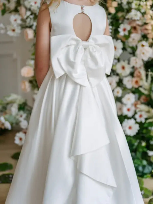 Penelope Off White Communion Dress shown from the back with a large bow and floral backdrop.