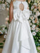 Penelope Off White Communion Dress shown from the back with a large bow and floral backdrop.