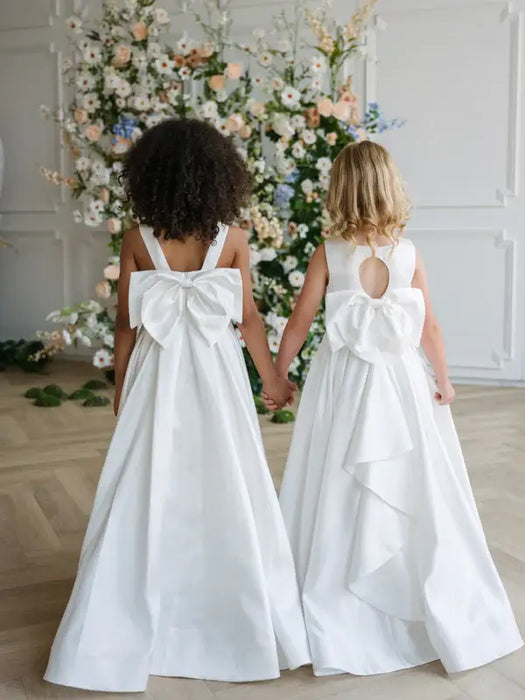 Teter Warm Penelope off-white communion dresses with large bows, worn by two girls holding hands indoors.
