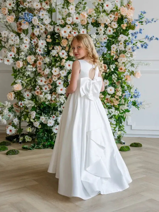 Teter Warm Penelope Off-White Communion Dress on girl with bow back, standing by floral backdrop.