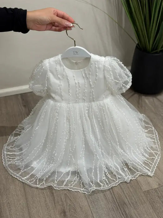 Teter Warm Perla off white sequin baptism dress displayed on a hanger, held above wood flooring.