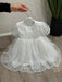 Teter Warm Perla off white sequin baptism dress displayed on a hanger, held above wood flooring.