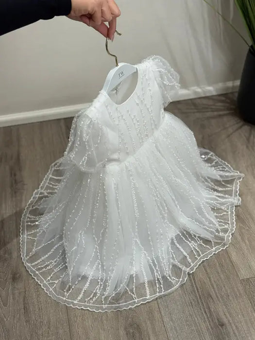 Teter Warm Perla off-white sequin baptism dress displayed on a hanger over a wood floor.