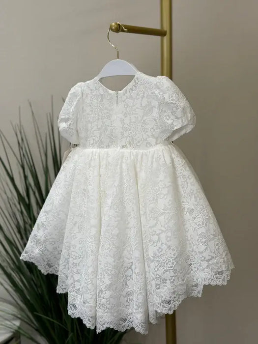 Warm Precious Lace Baptism Dress displayed on a hanger in a softly lit room.