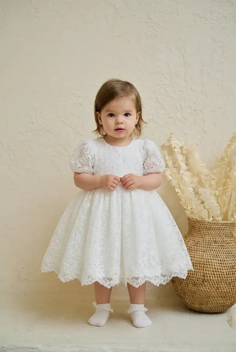 Precious Lace Baptism Dress shown on a toddler standing indoors near decorative wicker basket.
