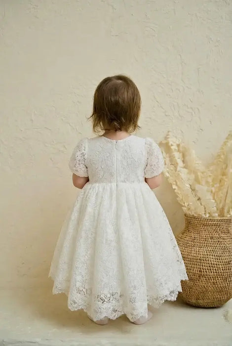 Precious Lace Baptism Dress shown on a toddler standing indoors near a wicker basket.