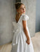Teter Warm Quinn Satin Communion Dress with lace sleeves and large back bow, worn by a girl indoors.