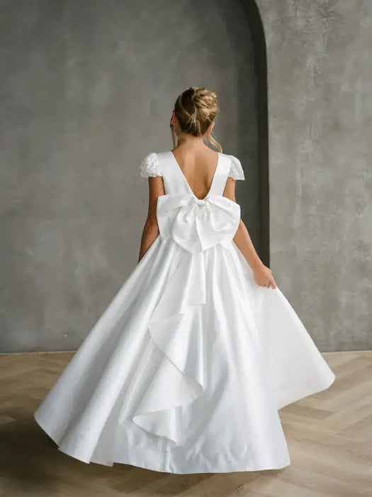 Teter Warm Quinn satin communion dress with large back bow shown on a girl in an elegant indoor setting.