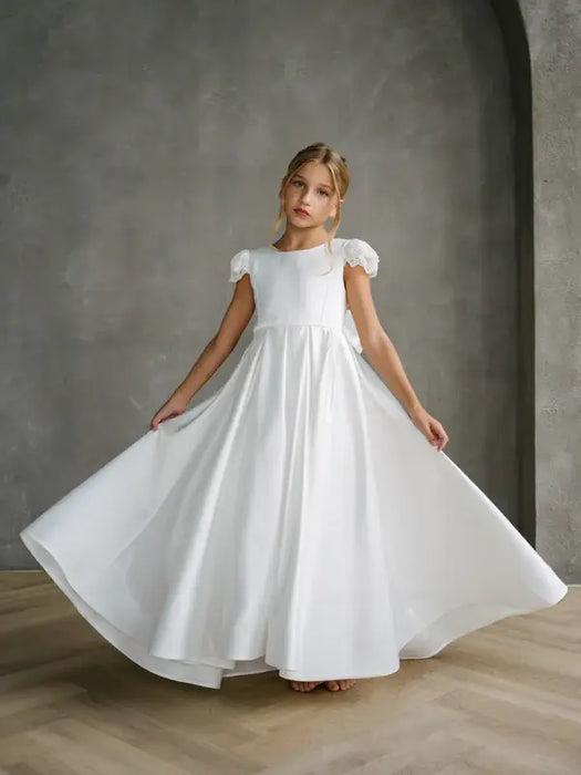 Quinn Satin Communion Dress shown on a young girl standing indoors, holding out the skirt.