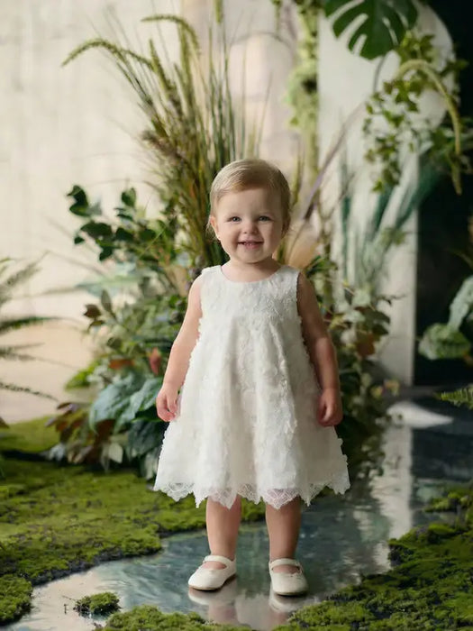 Teter Warm Rosalyn off-white floral baptism dress worn by a toddler standing on a reflective surface with greenery.