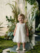 Teter Warm Rosalyn off-white floral baptism dress worn by a toddler standing on a reflective surface with greenery.