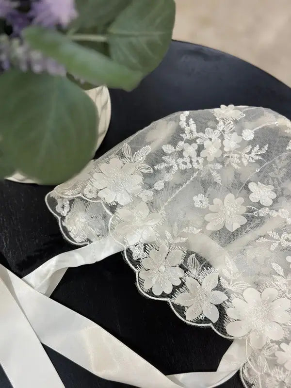 Teter Warm scalloped edge baby bonnet with floral lace, displayed on a black surface near a plant.