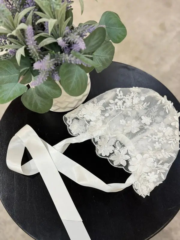 Teter Warm scalloped edge baby bonnet with floral lace detail displayed on a black table beside a potted plant.