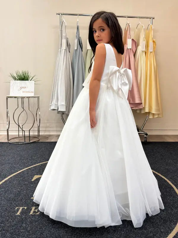 Teter Warm Shelly Satin Bow Communion Dress on a girl standing in a boutique with pastel dresses in the background.