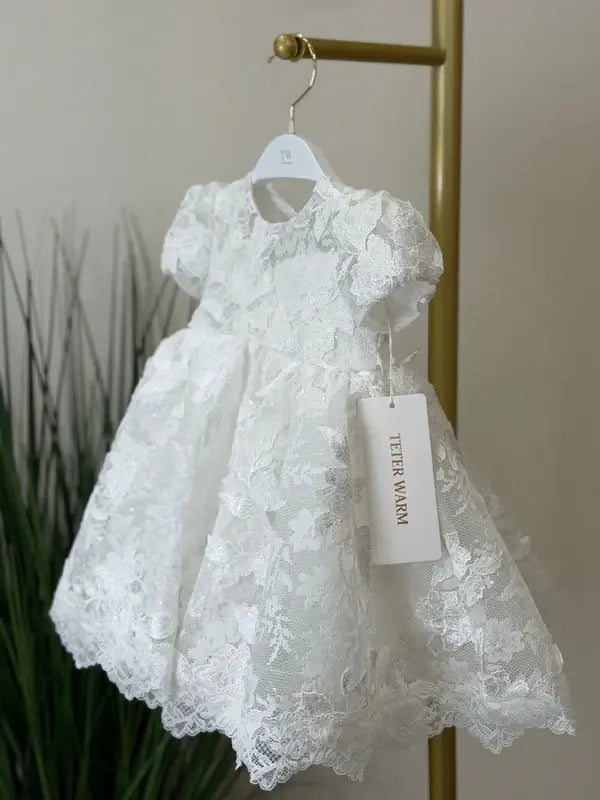 Teter Warm off-white floral lace baptism dress with puff sleeves shown hanging on a rack.