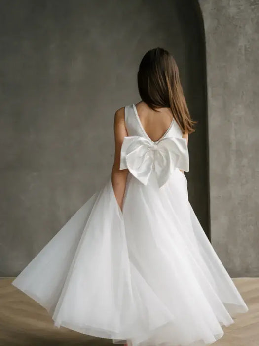 Teter Warm Sylvia Satin Pearl Communion Dress shown on a girl with a large bow in a minimal studio setting.