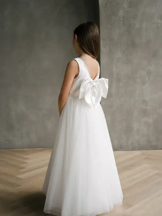 Sylvia Satin Pearl Communion Dress shown on a young girl with a large back bow, standing indoors.