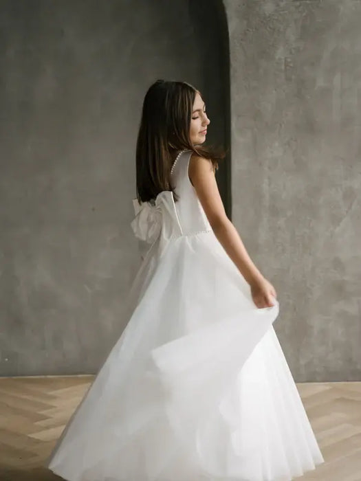 Sylvia Satin Pearl Communion Dress shown on a girl standing indoors with a tulle skirt and bow back.