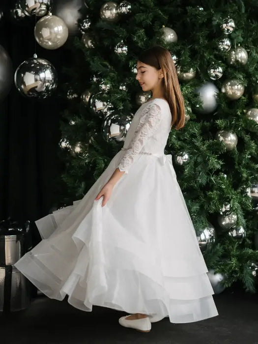 Teter Warm Taula Lace Communion Dress on a girl standing by a decorated Christmas tree with ornaments.