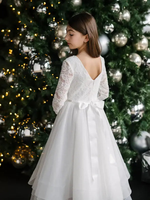 Teter Warm Taula lace communion dress with satin bow, shown on a young girl near a decorated Christmas tree.