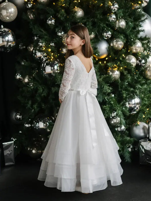Teter Warm Taula Lace Communion Dress shown on a girl standing in front of a decorated Christmas tree.
