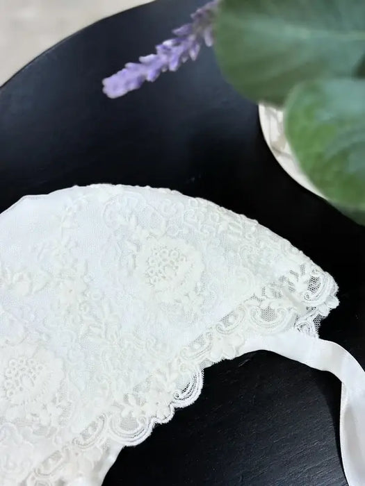 Teter Warm Ultra Soft Baby Bonnet in white lace displayed on a black tabletop near a plant.