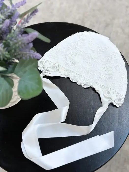 Teter Warm Ultra Soft Baby Bonnet with lace detail displayed on a black table beside a plant.