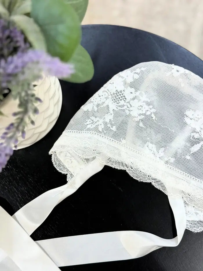 Valeria Lace Bonnet white baby bonnet with floral lace detail displayed on a dark table beside a vase of flowers.