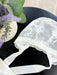 Valeria Lace Bonnet white baby bonnet with floral lace detail displayed on a dark table beside a vase of flowers.