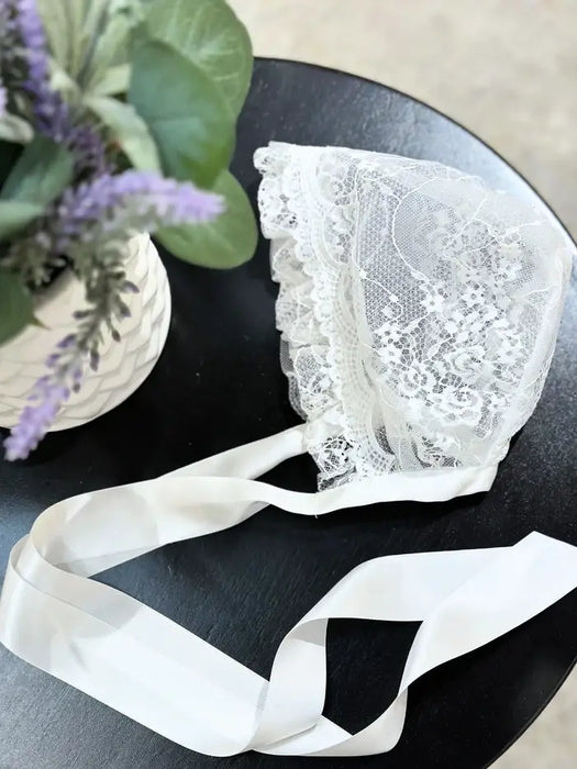 Teter Warm vintage baby bonnet with white lace and ribbon displayed on a round black table.