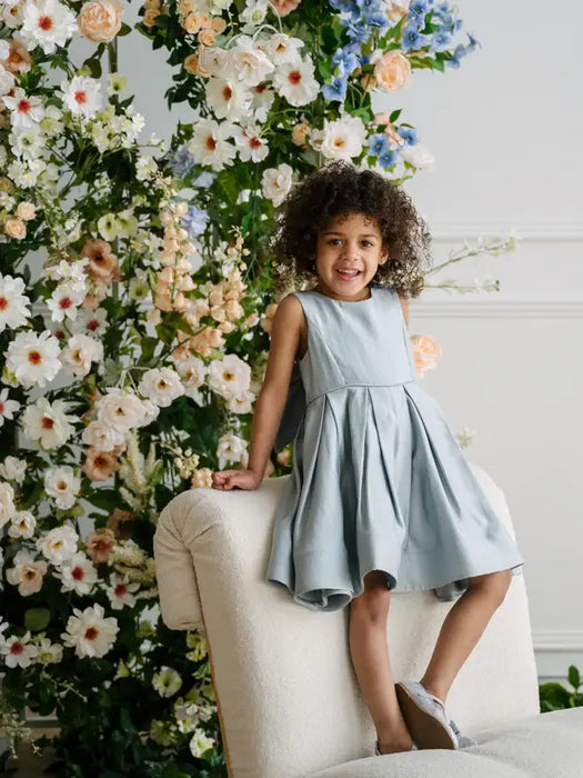 Teter Warm Zelda blue party dress worn by a smiling child sitting on a chair with floral backdrop.