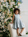 Teter Warm Zelda blue party dress worn by a smiling child sitting on a chair with floral backdrop.