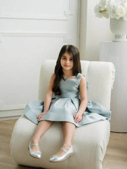 Teter Warm Zelda blue party dress on a young girl seated on a modern cream chair in a bright room.