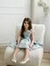 Teter Warm Zelda blue party dress on a young girl seated on a modern cream chair in a bright room.