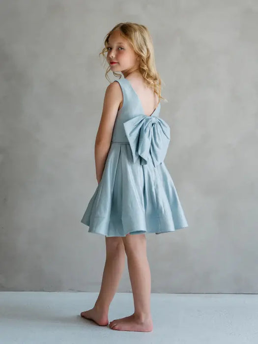 Teter Warm Zelda blue party dress with large back bow, shown on a young girl standing barefoot indoors.