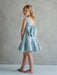 Teter Warm Zelda blue party dress with large back bow, shown on a young girl standing barefoot indoors.