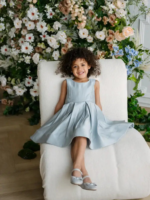 Teter Warm Zelda Blue Party Dress worn by a smiling girl sitting on a white chair with floral background.