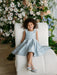 Teter Warm Zelda Blue Party Dress worn by a smiling girl sitting on a white chair with floral background.